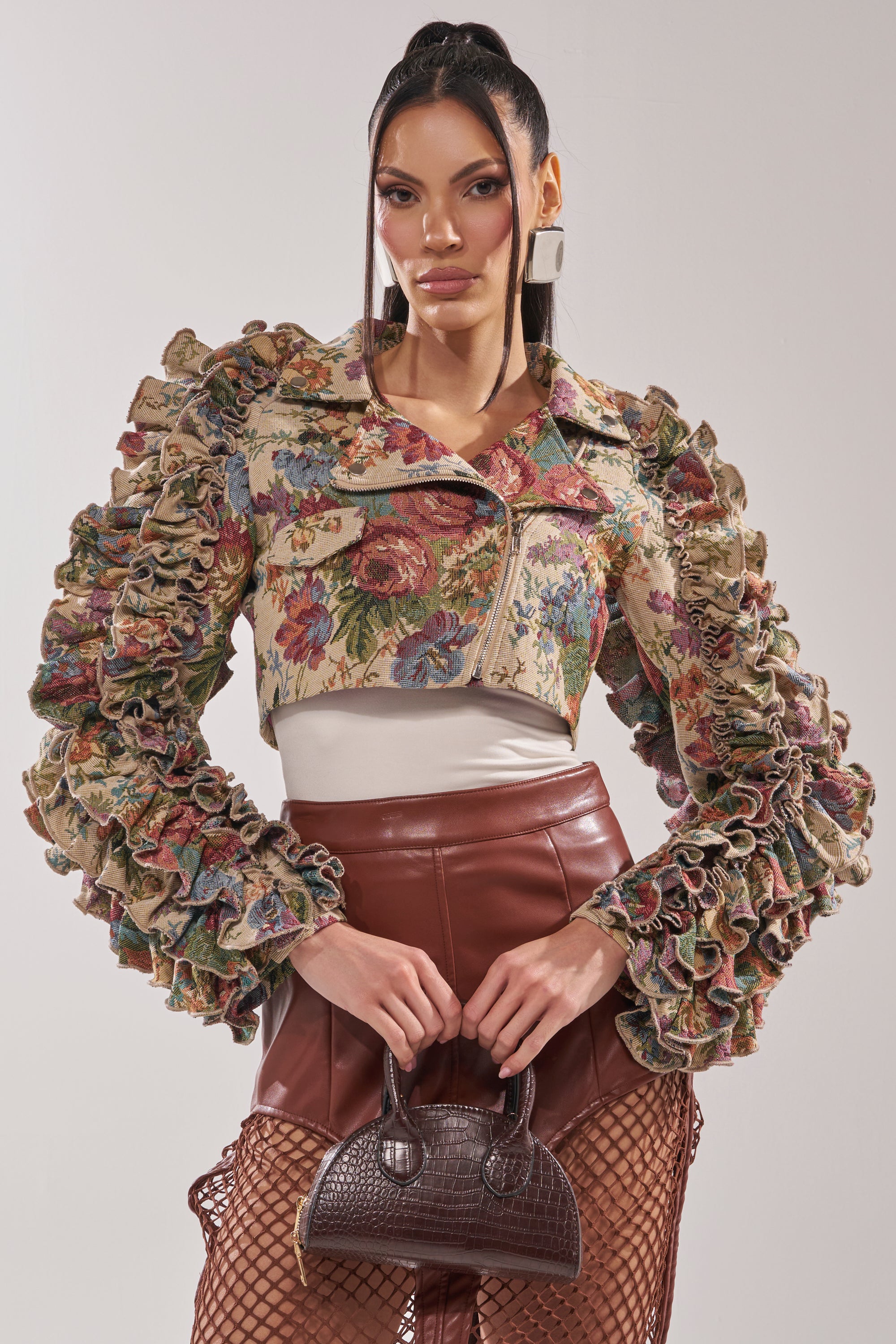 A woman poses in the MAKE IT LOOK EASY TAPESTRY MOTO jacket with ruffled sleeves, a cream top, brown faux leather shorts, fishnet stockings, and a small dark brown handbag against a light background. 1