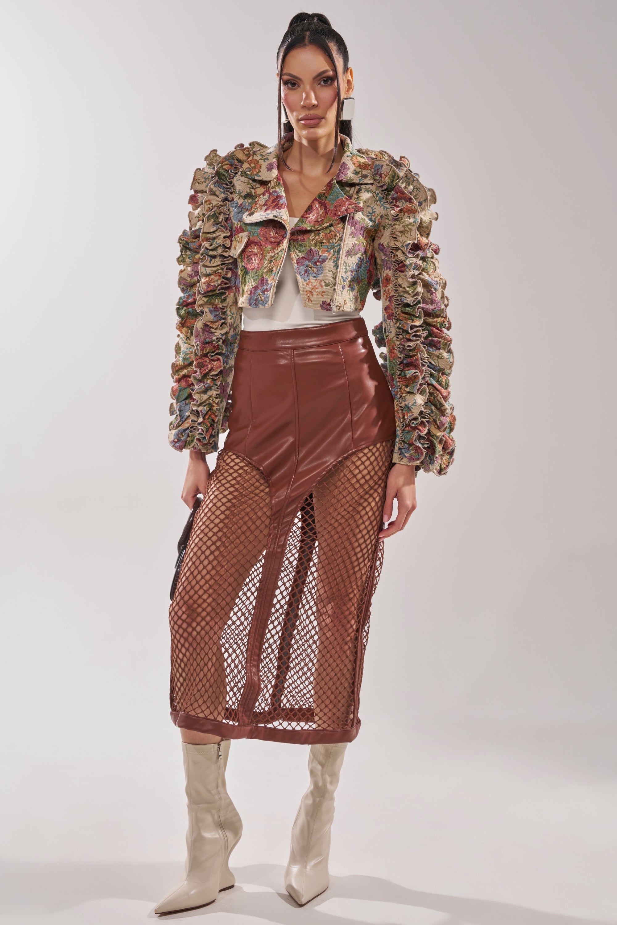 A woman stands against a plain background wearing the MAKE IT LOOK EASY TAPESTRY MOTO jacket with ruffled sleeves, a white top, a brown leather skirt with sheer fishnet panels, and off-white pointed boots. 9