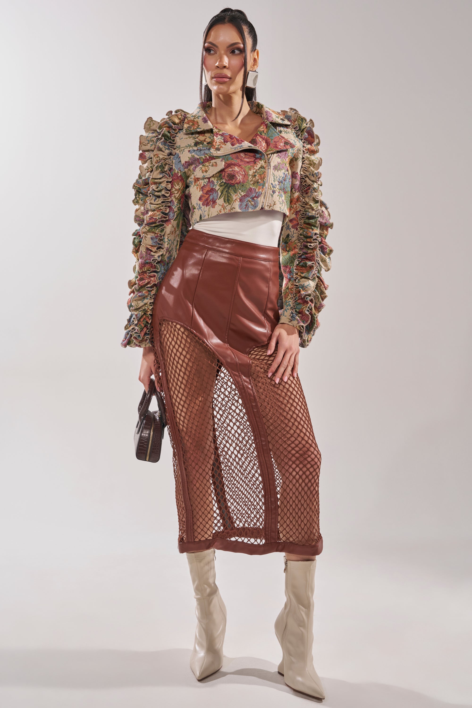 A woman poses in the MAKE IT LOOK EASY TAPESTRY MOTO jacket, a brown faux leather skirt with mesh panels, white boots, and a small brown handbag against a plain background. 10