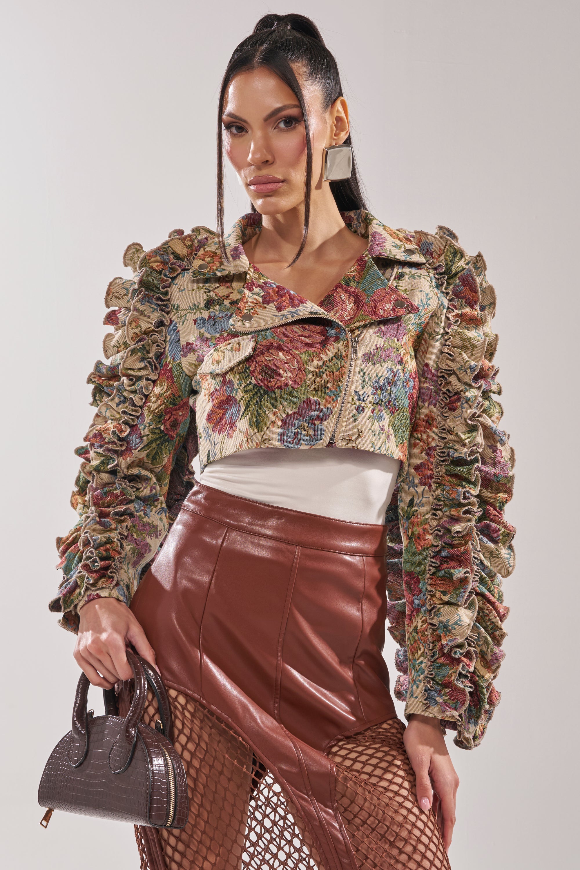 A woman models the MAKE IT LOOK EASY TAPESTRY MOTO jacket over a white top, styled with a brown faux leather skirt, fishnet stockings, a small handbag, and straight hair. 0