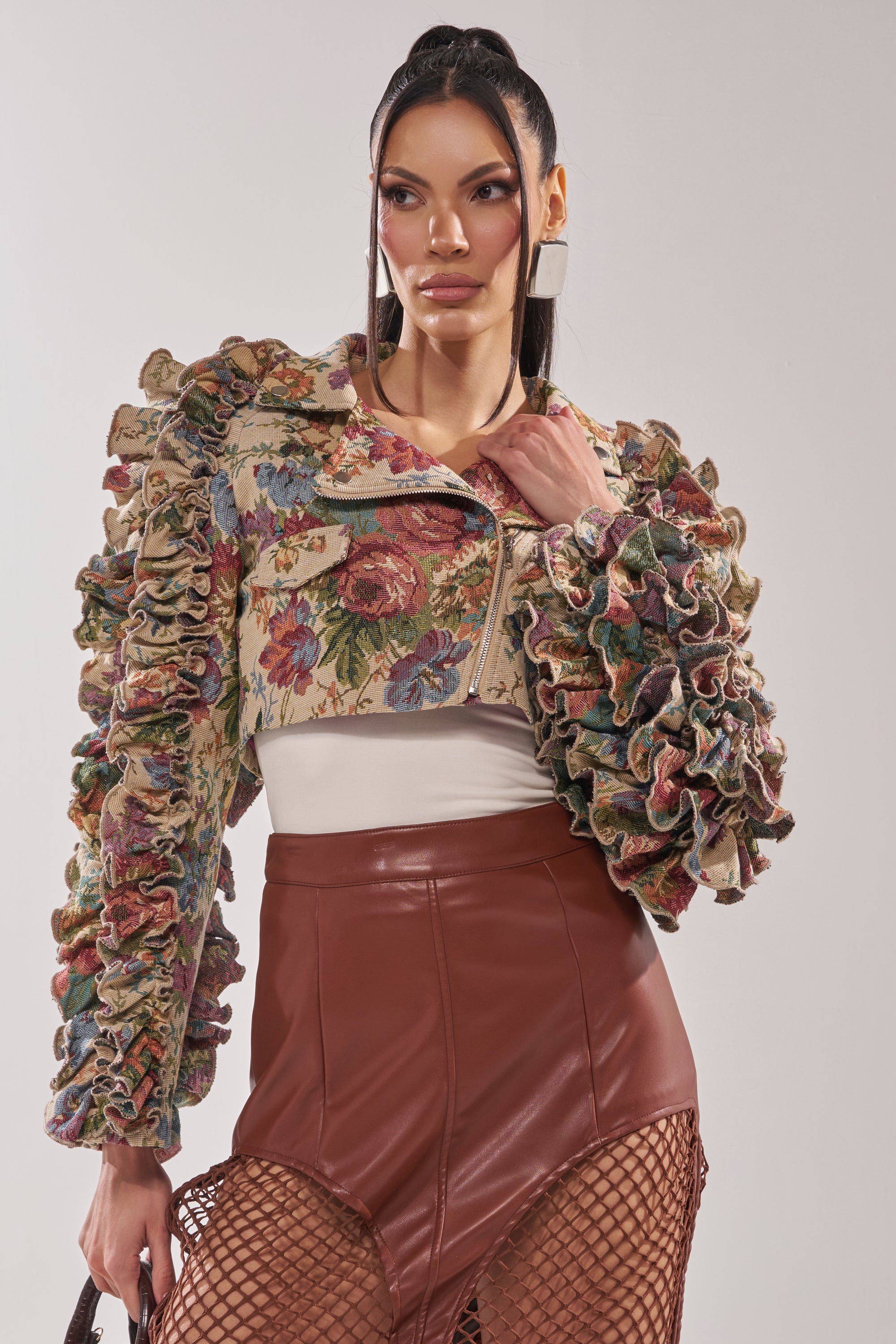 A woman models the MAKE IT LOOK EASY TAPESTRY MOTO jacket with ruffled sleeves over a white top and a high-waisted brown faux leather skirt with a fishnet layer. Her long dark hair is styled in a high ponytail. 2