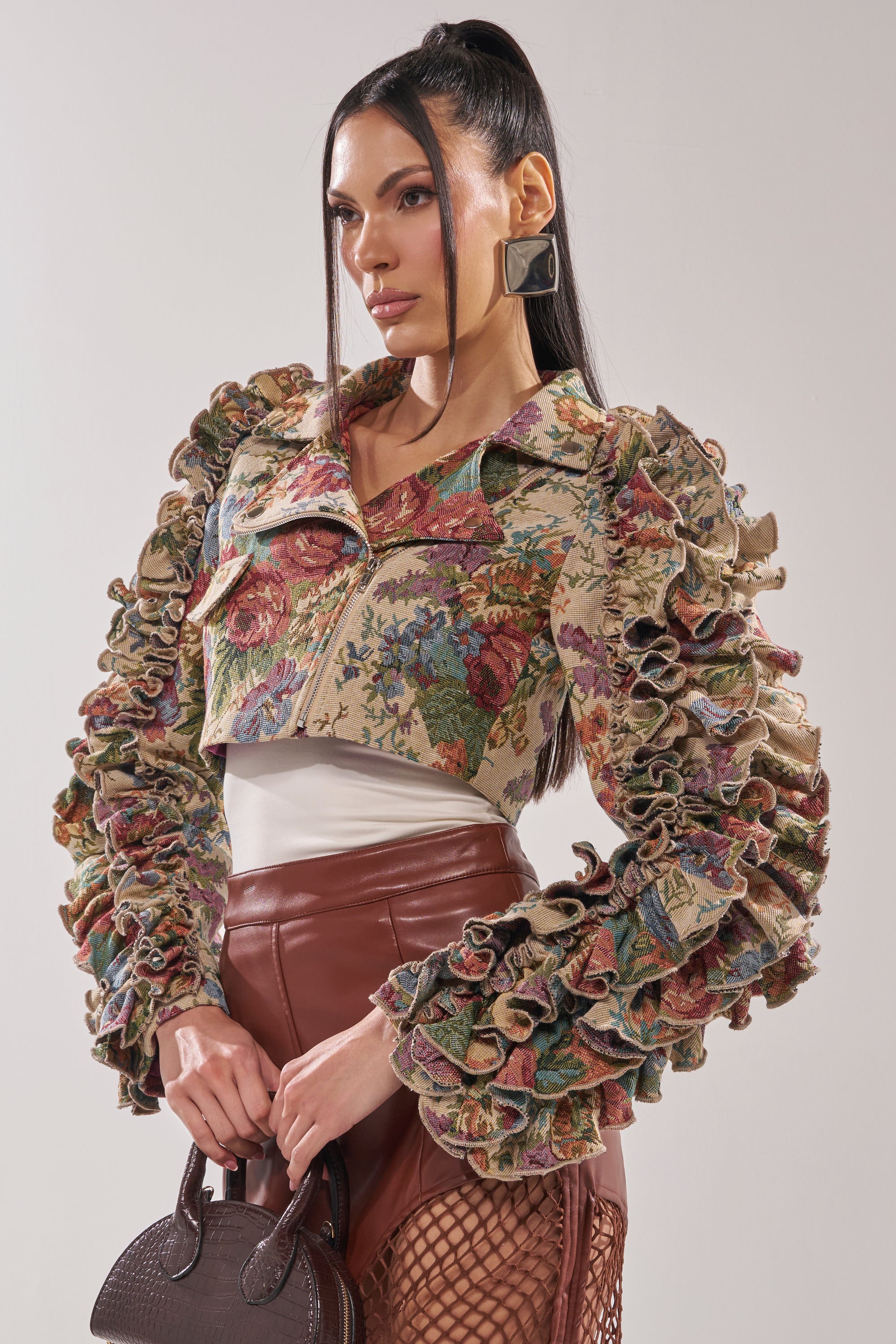 A woman with long dark hair in a high ponytail wears the MAKE IT LOOK EASY TAPESTRY MOTO jacket, brown leather pants with mesh cutouts, square earrings, and holds a textured brown handbag against a neutral background. 3