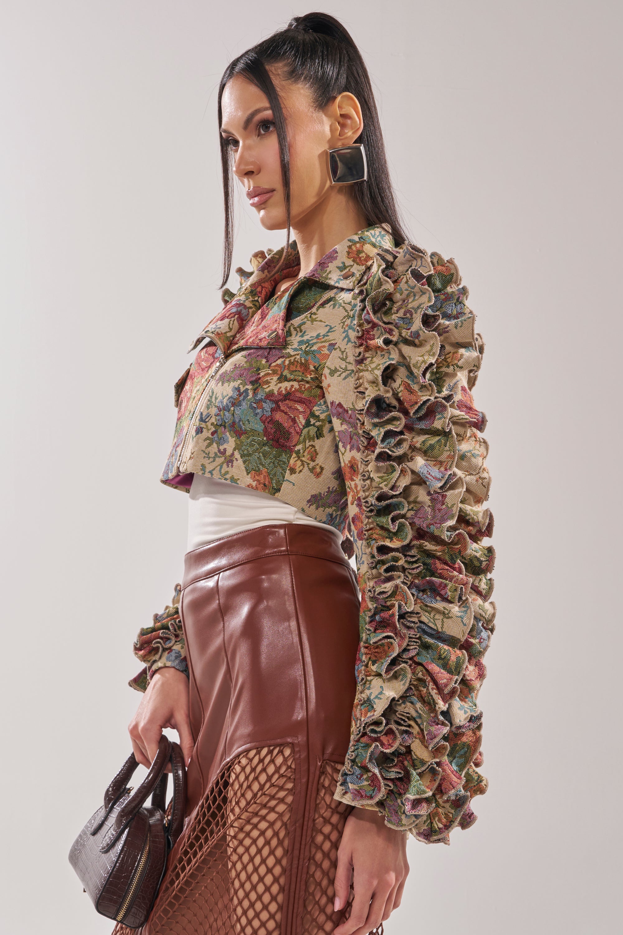 A woman wears the MAKE IT LOOK EASY TAPESTRY MOTO jacket with ruffled sleeves, a brown faux leather skirt featuring mesh panels, and carries a textured handbag. She styles her long dark hair in a high ponytail with large square earrings. 4