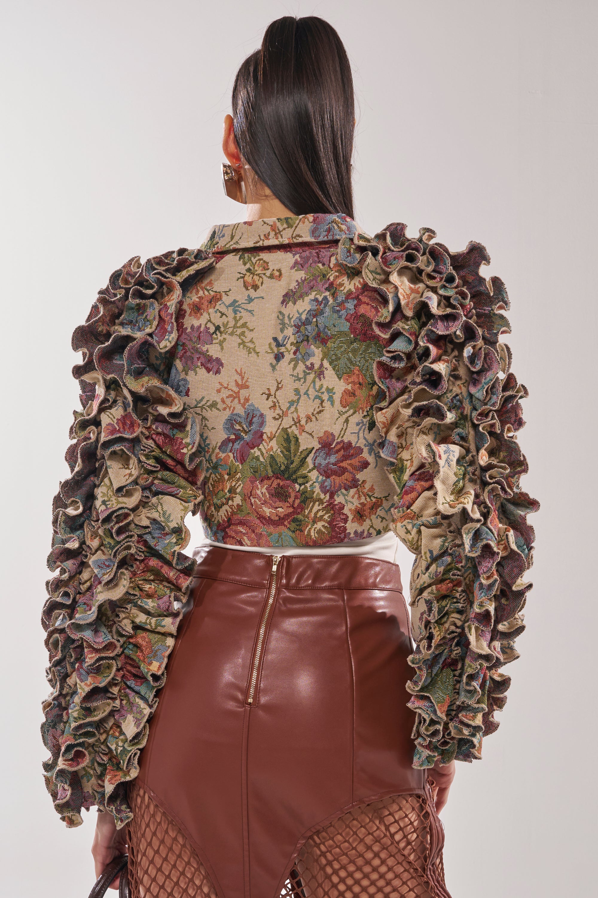 A woman with long dark hair in a ponytail, seen from the back against a plain background, wears the MAKE IT LOOK EASY TAPESTRY MOTO jacket with ruffled sleeves and a high-waisted brown faux leather skirt with a zipper. 5