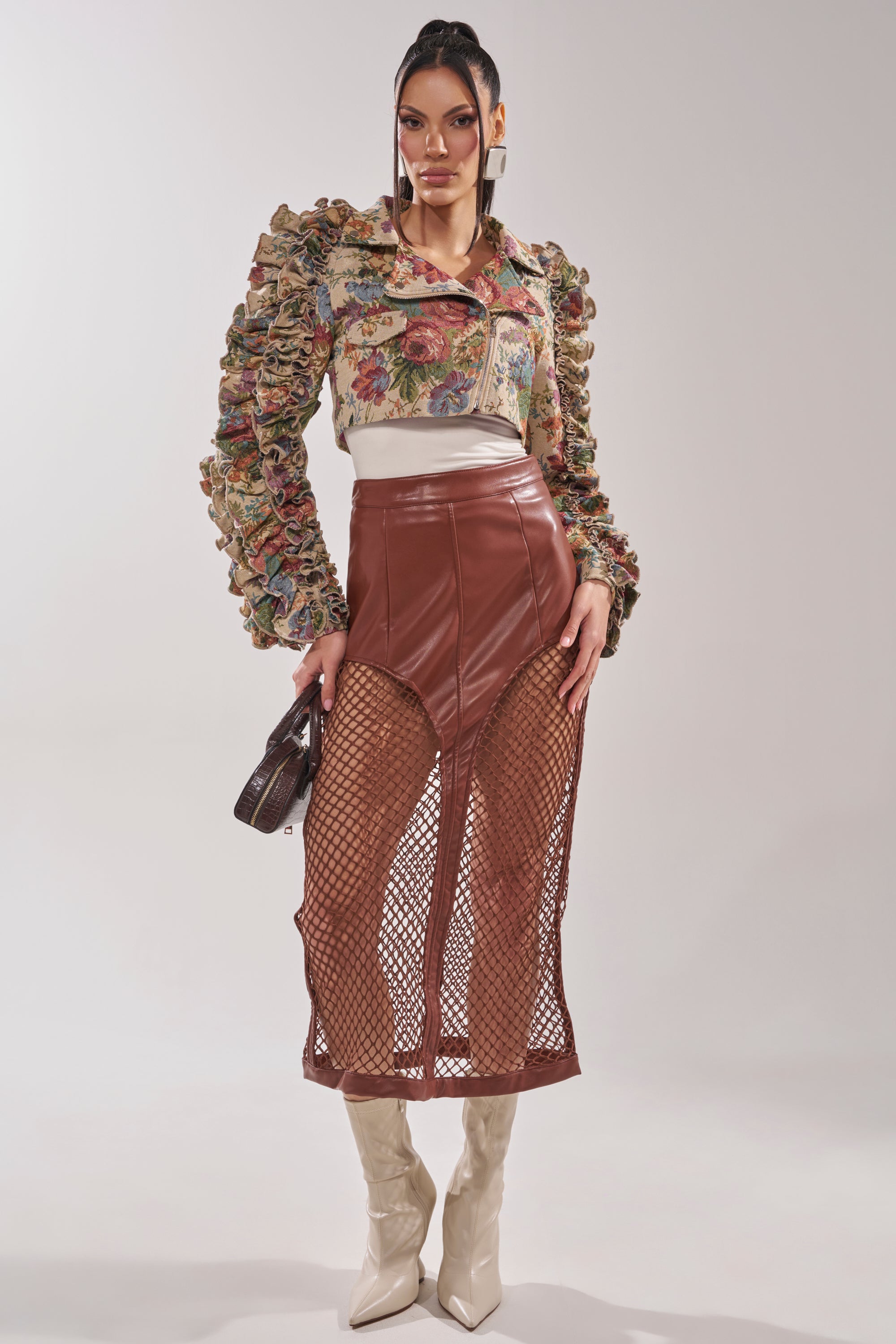 A woman wears the MAKE IT LOOK EASY TAPESTRY MOTO jacket over a floral ruffled blouse, high-waisted brown leather skirt with mesh panels, cream knee-high boots, and carries a small dark handbag against a light background. 6