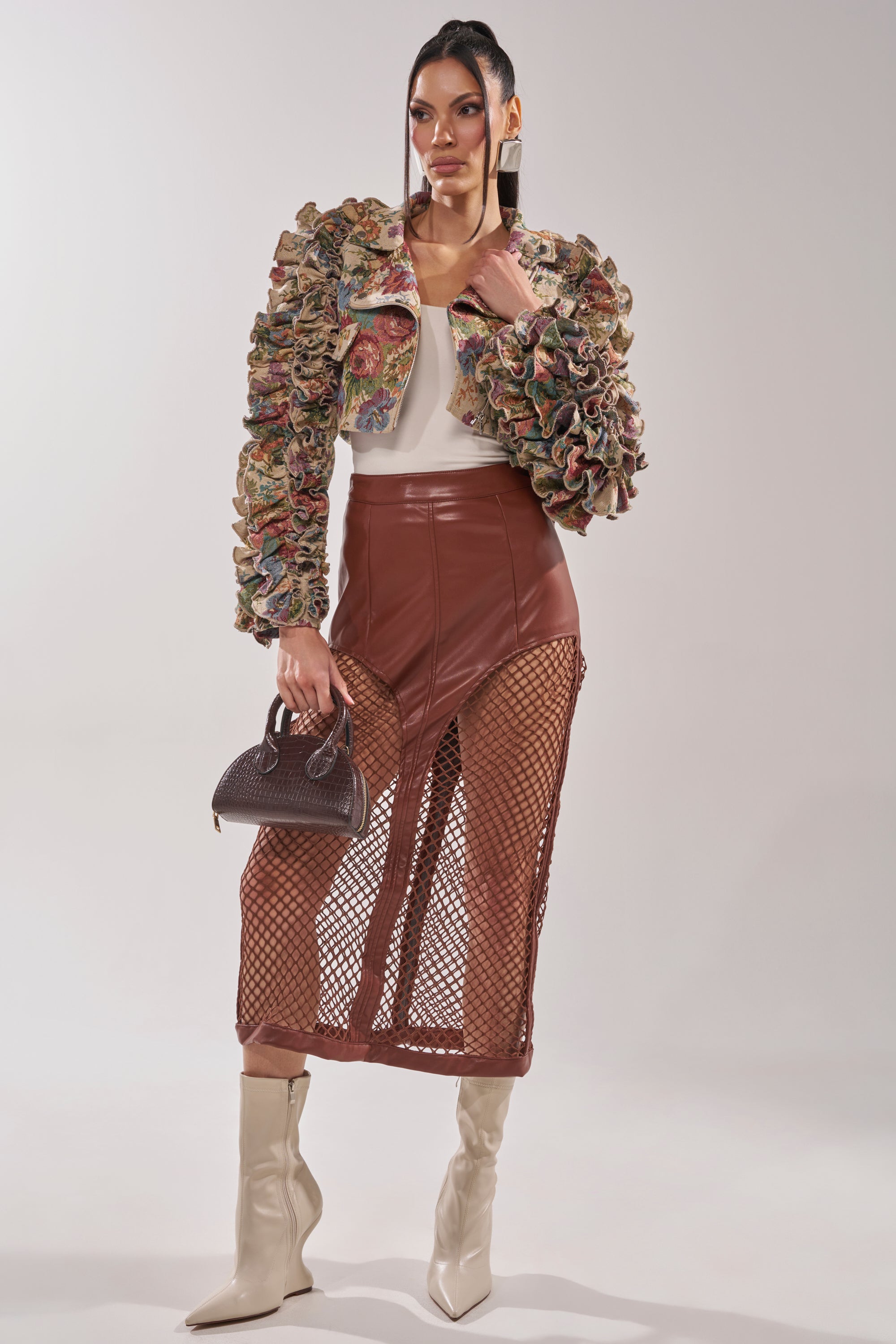 A woman poses in the MAKE IT LOOK EASY TAPESTRY MOTO jacket with ruffled details, paired with a white top, brown leather skirt with fishnet panels, cream boots, small brown handbag, sleek straight hair, and statement earrings. 7
