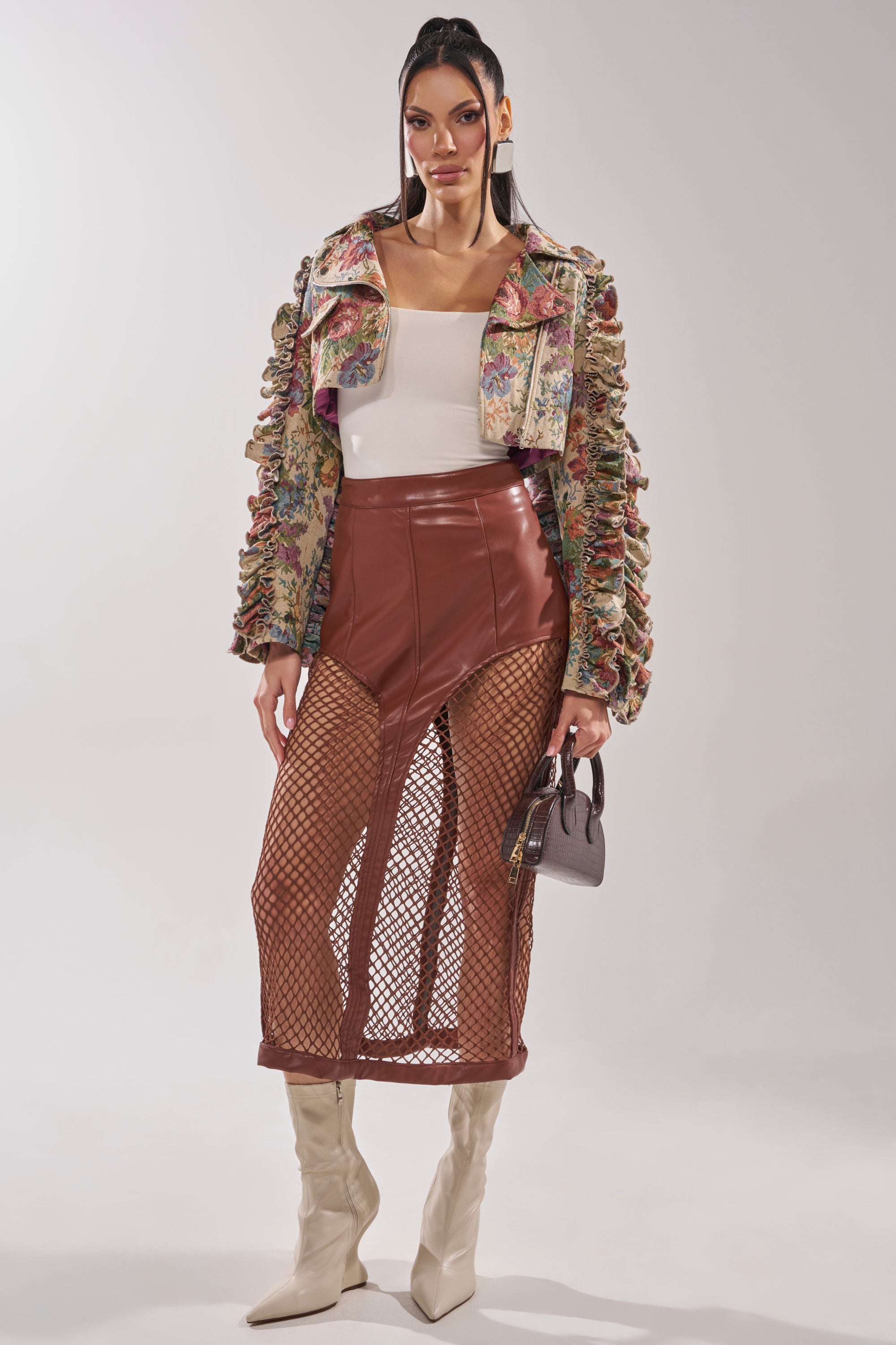 A woman poses in the MAKE IT LOOK EASY TAPESTRY MOTO jacket, styled with a white top, high-waisted brown leather skirt with mesh panels, knee-high white boots, and a small gray handbag against a plain background. 8