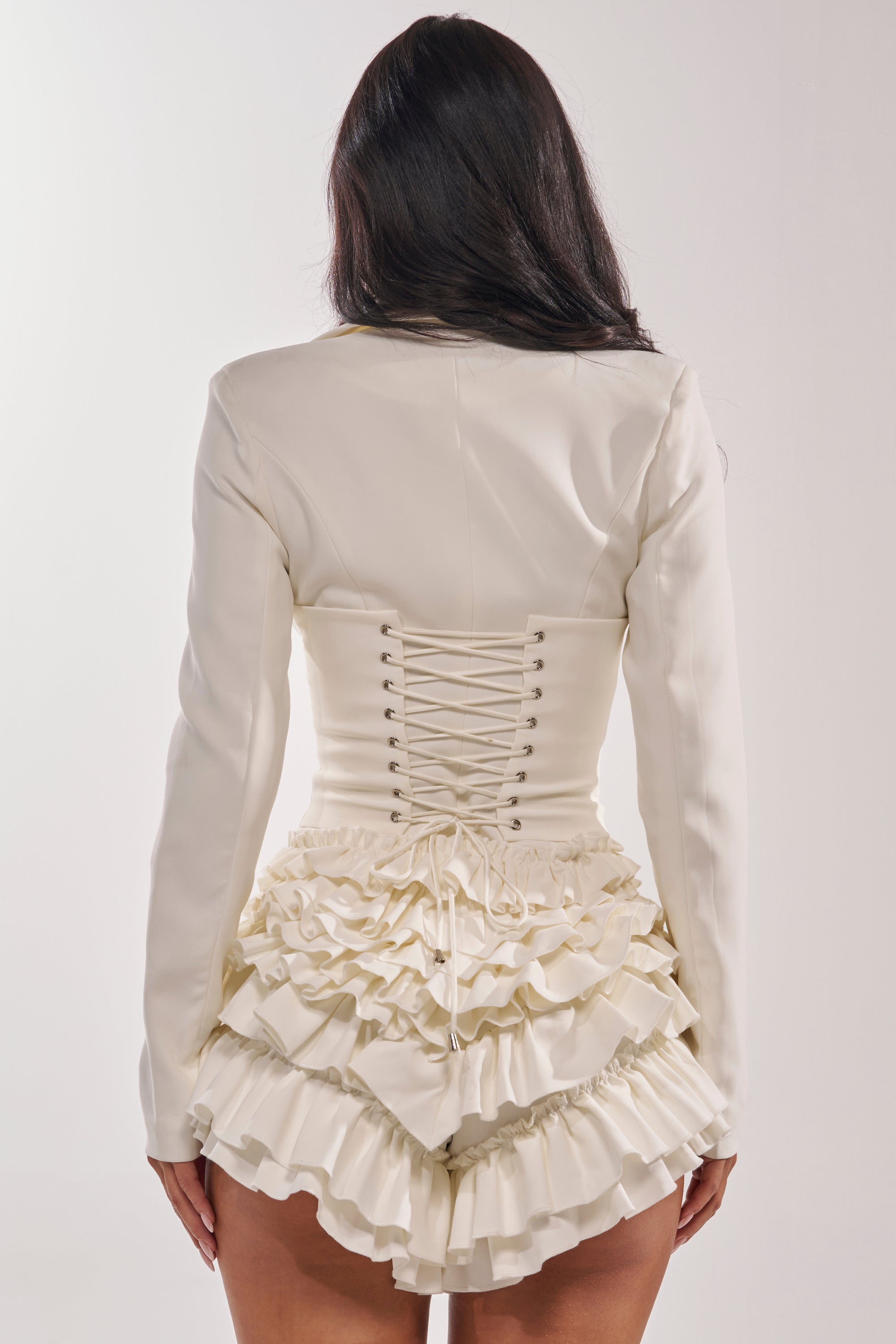A woman with long dark hair models the STRICTLY BUSINESS BLAZER ROMPER IN WHITE, which features a lace-up corset back and layered ruffle hem, posing with her back to the camera against a plain light background. 4