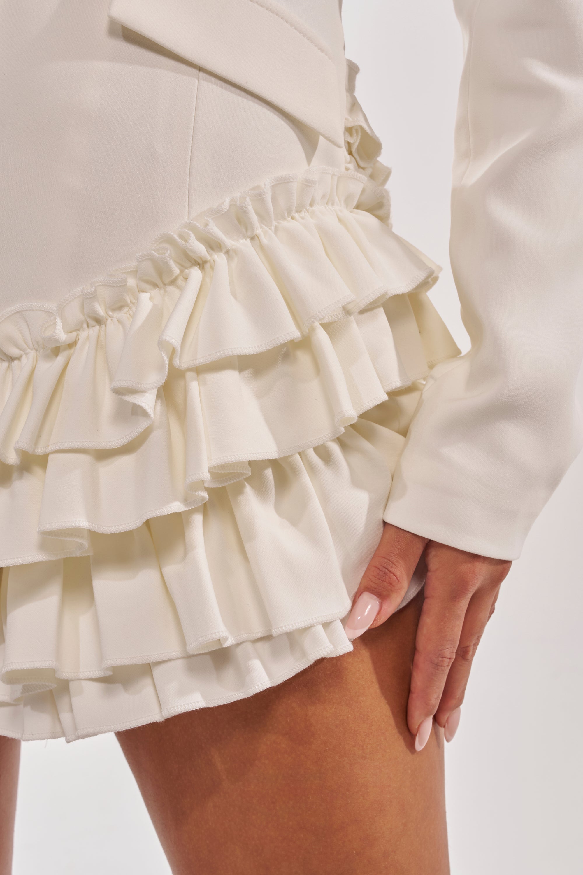 A close-up of a person wearing the STRICTLY BUSINESS BLAZER ROMPER IN WHITE, featuring layered skirt ruffles and elegant fabric. Their manicured hand on their thigh highlights its chic style—ideal for bridal or cocktail occasions. 6