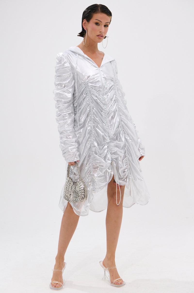 A woman stands against a white background wearing the WAYS TO GO METALLIC ADJUSTABLE DRESS IN WHITE, paired with a small silver handbag, clear high-heeled sandals, slicked-back hair, and large hoop earrings.