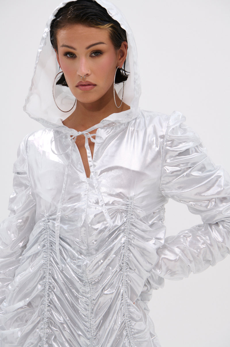 A person wears the WAYS TO GO METALLIC ADJUSTABLE DRESS IN WHITE, styled with a hooded jacket design and tie neckline, paired with large hoop earrings, posing confidently against a plain white background.