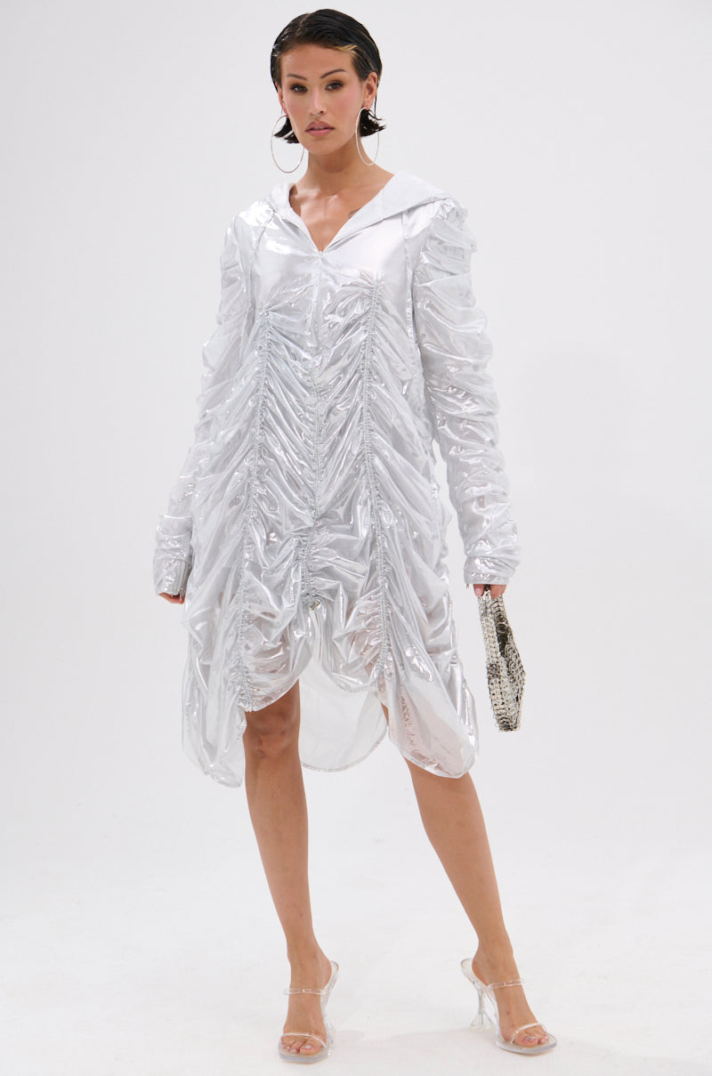 A woman poses against a plain white background wearing the WAYS TO GO METALLIC ADJUSTABLE DRESS IN WHITE with adjustable ruching, long sleeves, and a hood, holding a small textured clutch and clear high-heeled shoes.