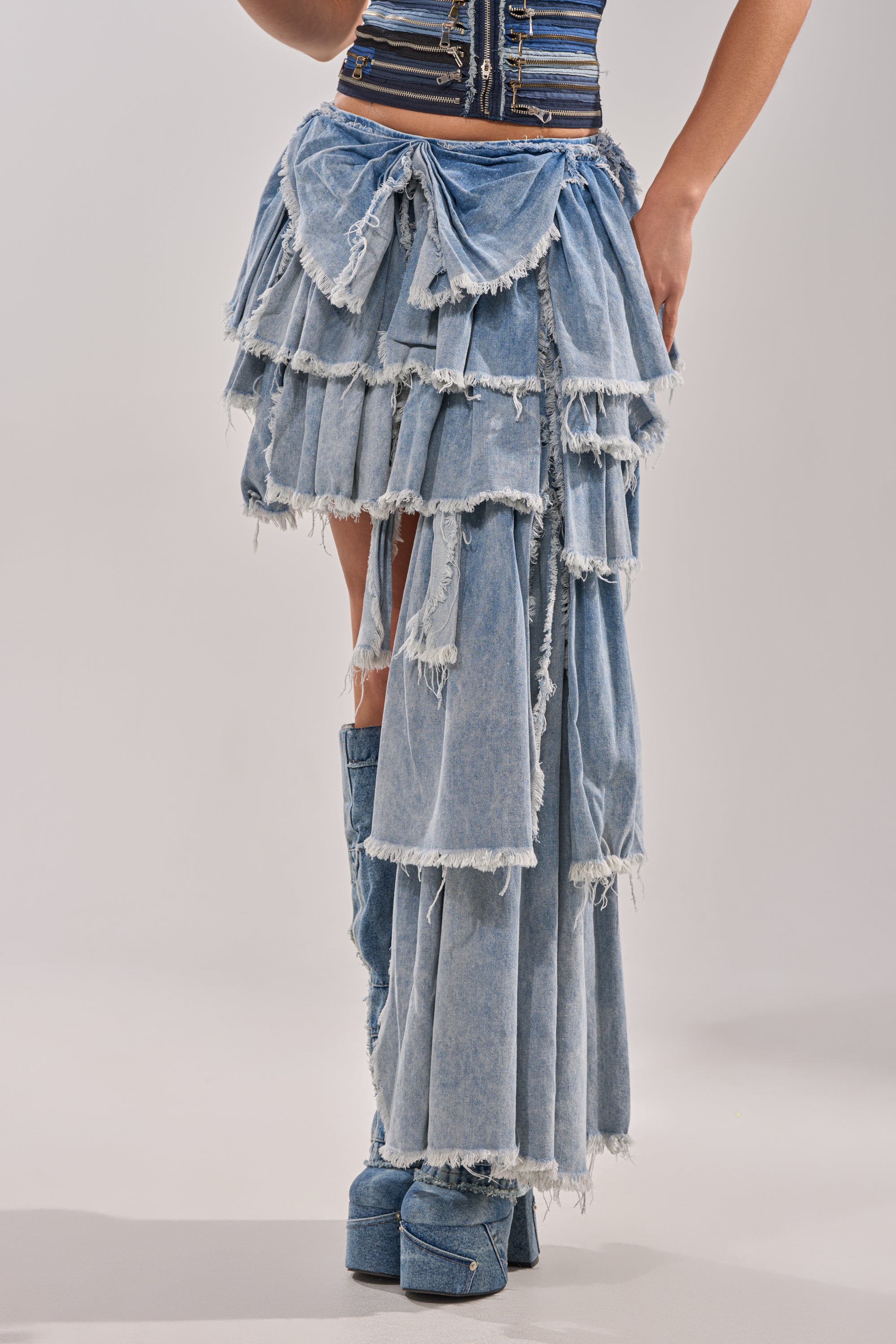 A person wearing the DISTRESSED TIERED ASYMMETRIC MIDI SKIRT with frayed edges, paired with matching denim platform boots and a striped fitted top for a textured, avant-garde look. 3