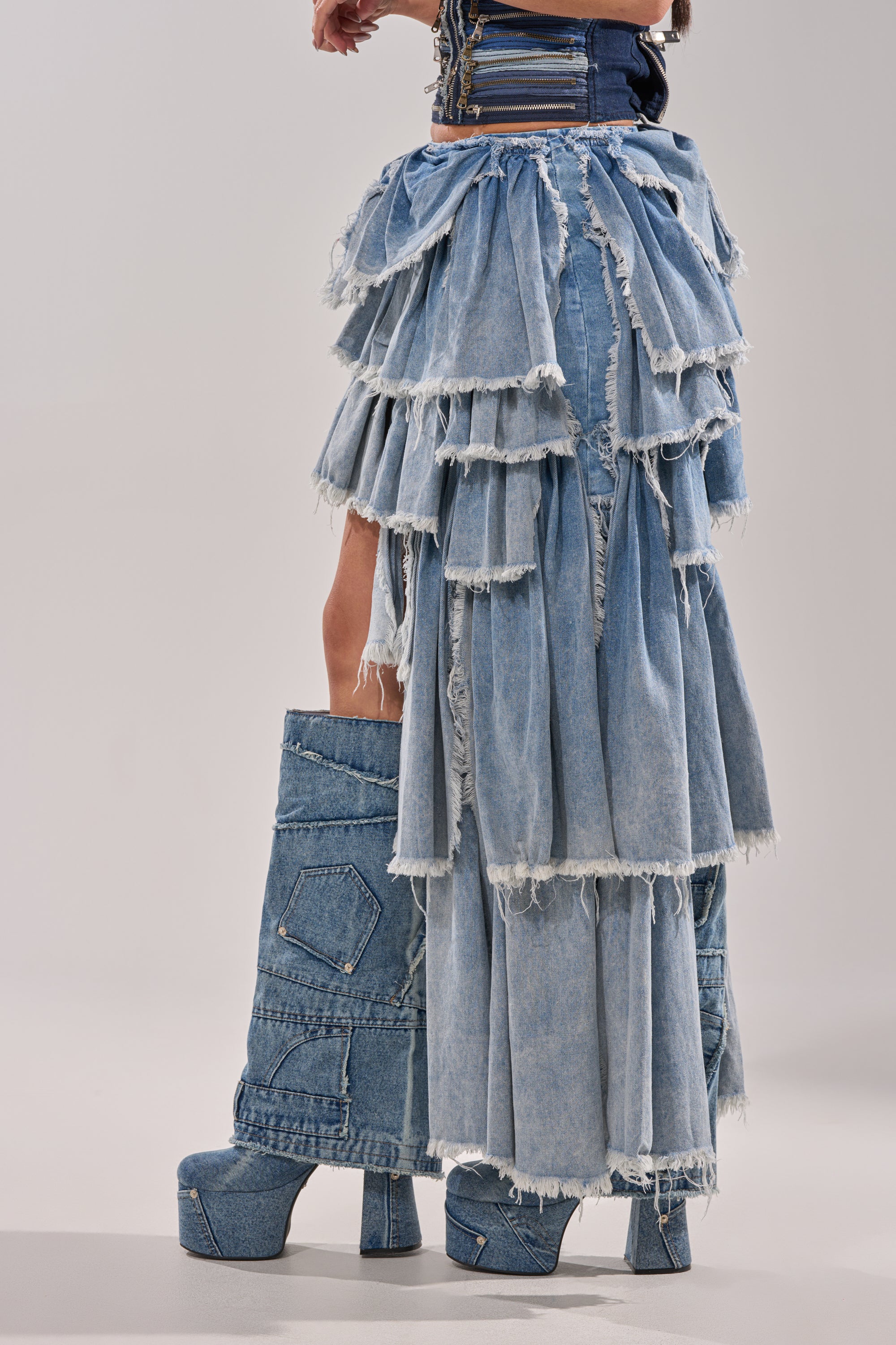 Wearing the DISTRESSED TIERED ASYMMETRIC MIDI SKIRT with ruffled denim layers and matching platform denim boots with visible seams and frayed edges, a person stands against a plain background. 4