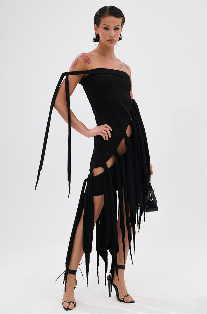 A person stands confidently in the black REVERIE MINI DRESS WITH TIES, showcasing its asymmetrical design, dramatic slits, and long hanging ties, paired with high heels and a small black handbag against a plain white background. 0