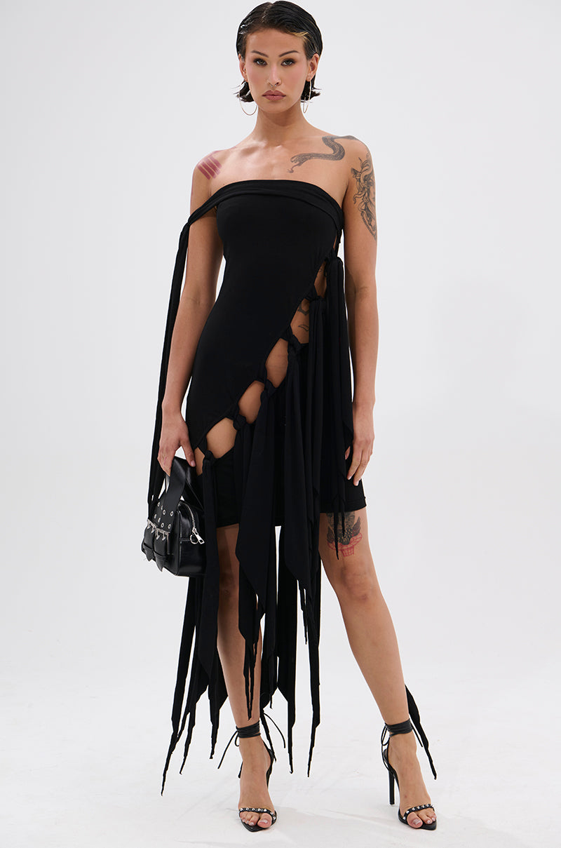 A woman stands confidently in a black REVERIE MINI DRESS WITH TIES with an asymmetrical cut, cutouts, and a handkerchief hem, styled with black heels, a small black handbag, and showing tattoos on her arm and shoulder. 1