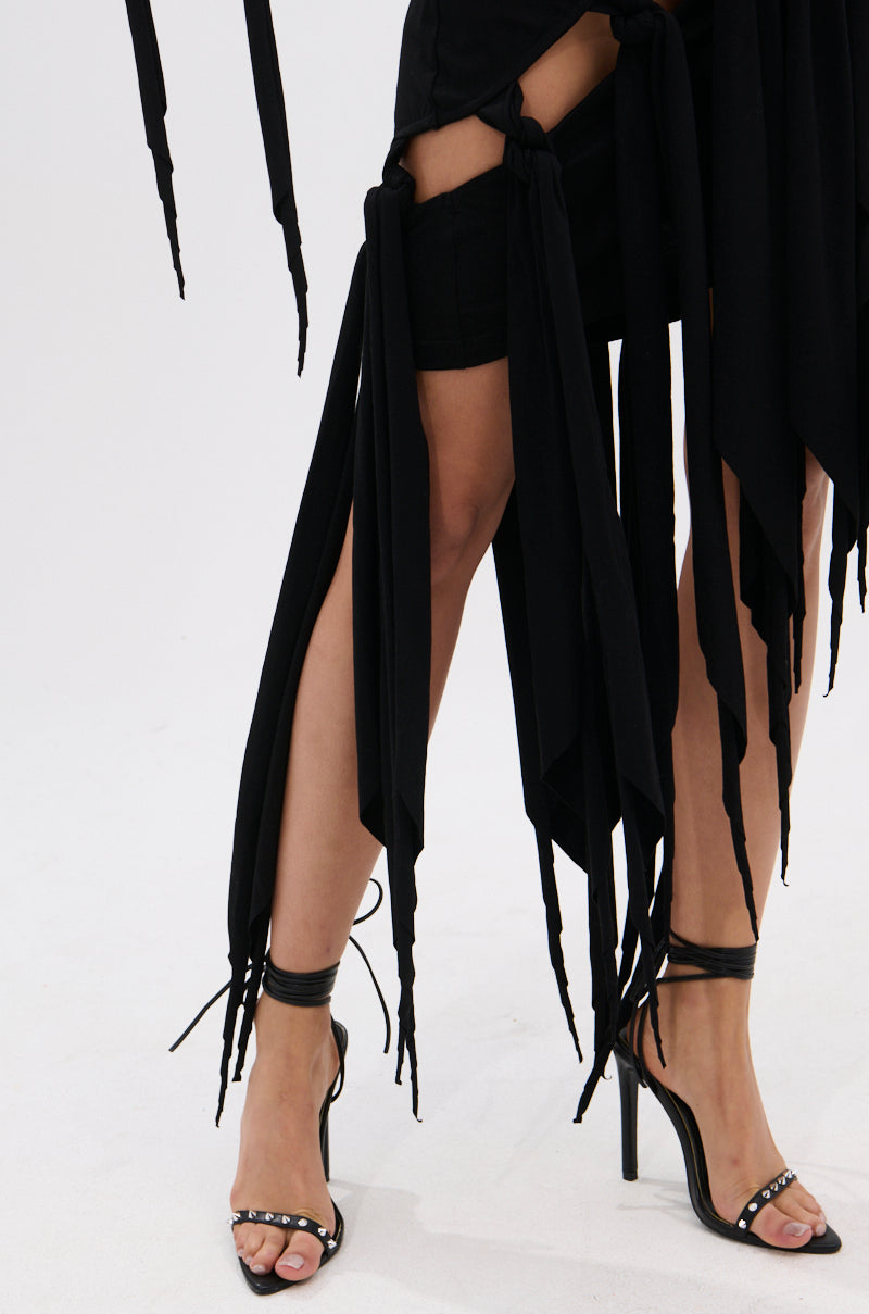 A person wearing the REVERIE MINI DRESS WITH TIES—a black skirt featuring a handkerchief hem and long, flowing ties—paired with strappy black high heels, stands on a light-colored floor. 5