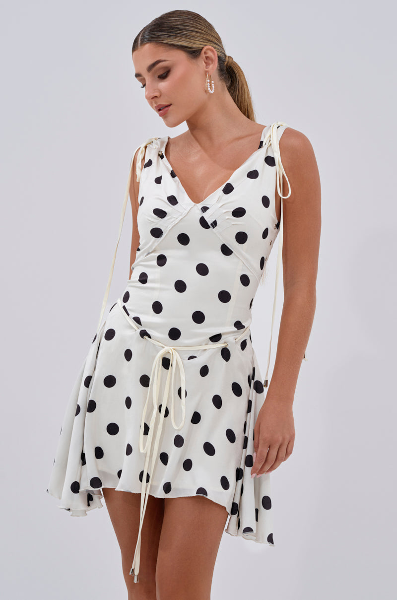 A woman wears the SWEET TALK POLKA DOT MINI DRESS, featuring a sleeveless A-line silhouette with tie straps and a flared skirt, standing against a plain light background.