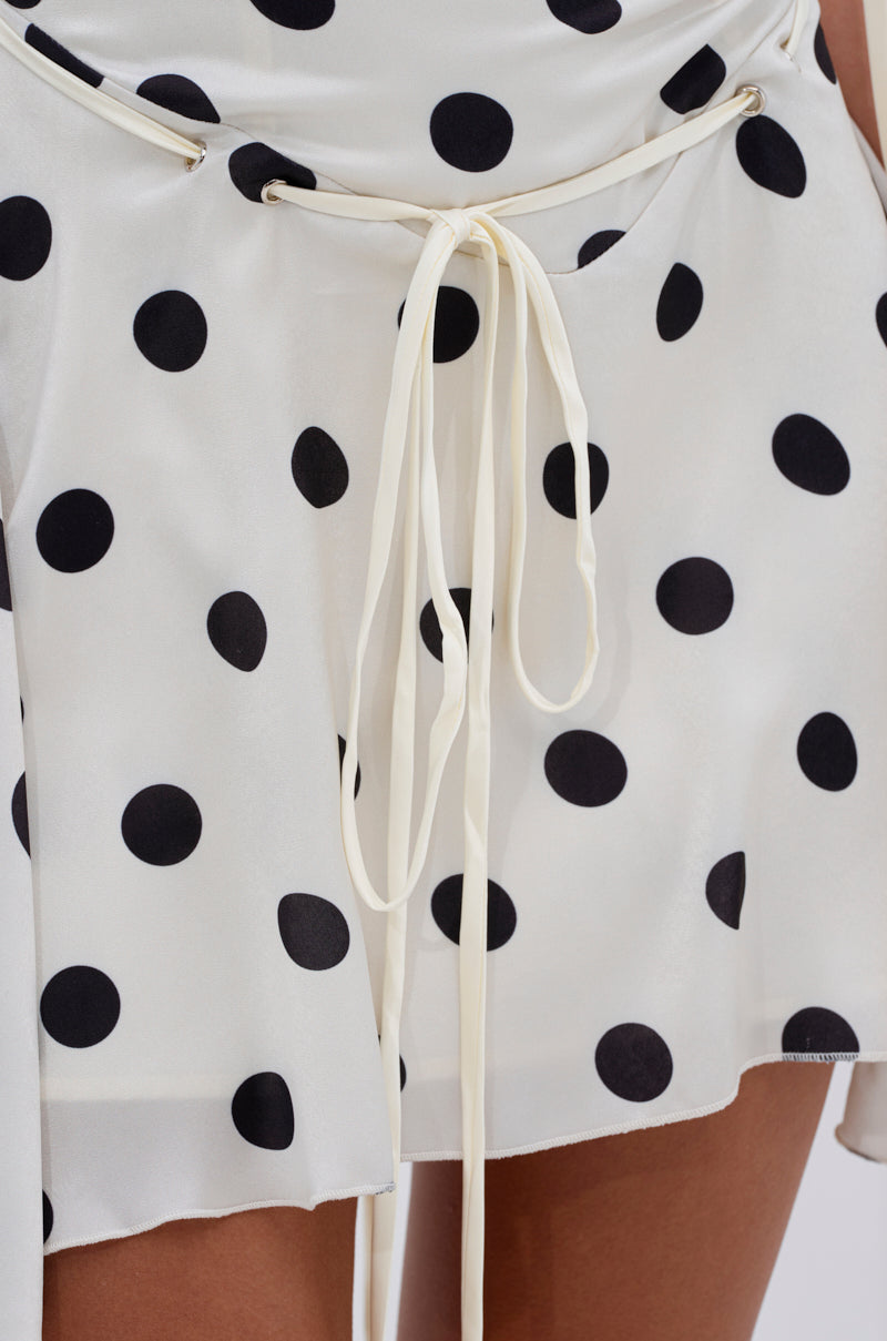 The SWEET TALK POLKA DOT MINI DRESS features large black spots, a thin white tie belt at the waist, and light, slightly sheer fabric with an asymmetrical hem—perfect for a playful and breezy look.