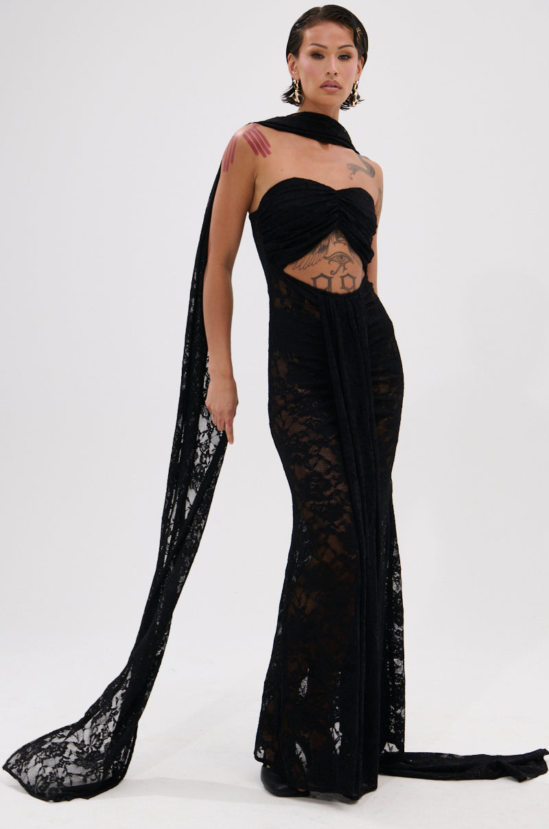A person stands confidently in the READY FOR A NIGHT OUT LACE MAXI DRESS, featuring sheer panels and a long matching scarf, showing off tattoos and styled with slicked-back hair and bold earrings.
