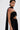 A person with short, slicked-back hair wears the READY FOR A NIGHT OUT LACE MAXI DRESS—a sheer, column-silhouette maxi styled with a scarf over one shoulder. Tattoos decorate their chest; bold red makeup pops on their left shoulder against a white background.