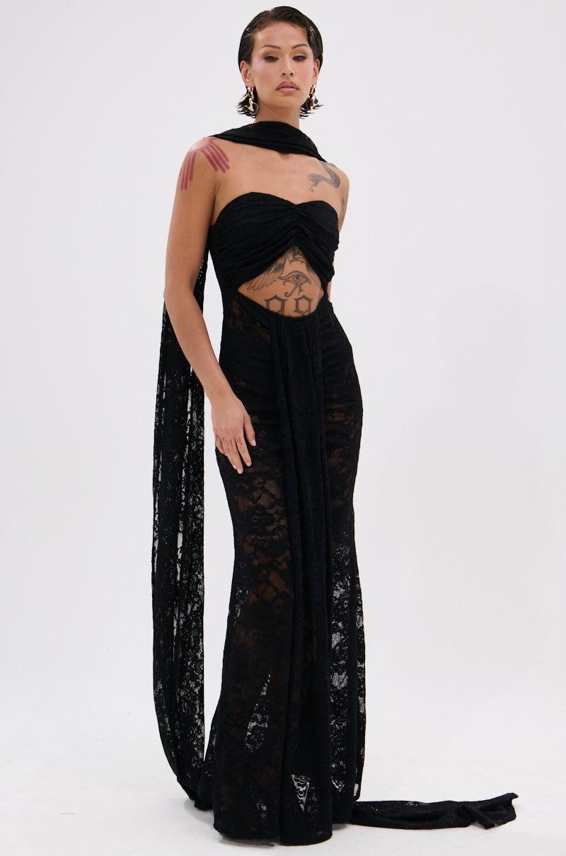 A person poses confidently in the READY FOR A NIGHT OUT LACE MAXI DRESS, a sheer strapless maxi with waist cutout and long matching scarf. They have chest tattoos, bold earrings, and slicked-back hair against a plain white background.
