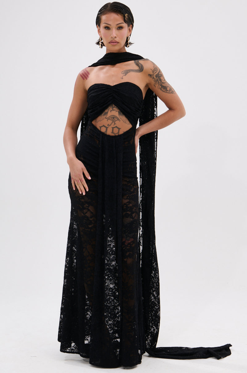 A woman poses confidently against a white background in the READY FOR A NIGHT OUT LACE MAXI DRESS, featuring a strapless design, sheer lace skirt, and cut-out midsection. She accessorizes with statement earrings and a long black scarf.