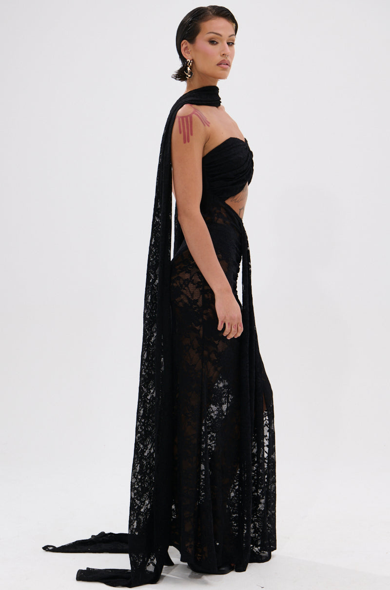 A woman stands sideways in the READY FOR A NIGHT OUT LACE MAXI DRESS, a strapless black sheer lace column gown with a matching scarf. Her slicked-back hair, bold dark earrings, and minimal, confident expression complete the look.