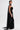 A woman stands sideways in the READY FOR A NIGHT OUT LACE MAXI DRESS, a strapless black sheer lace column gown with a matching scarf. Her slicked-back hair, bold dark earrings, and minimal, confident expression complete the look.