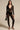 Against a neutral backdrop, someone wearing the "Wish You Were Me Lace Jumpsuit" with a plunging neckline exudes vintage romance. They accessorize with a small black handbag and high-heeled sandals, while their long hair is perfectly complemented by elegant earrings and a necklace.