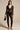 A woman radiates vintage romance in the WISH YOU WERE ME lace jumpsuit, featuring a sheer black design and plunging neckline. She complements the look with a small black handbag, high heels, and long straight hair, all against a simple beige backdrop enhancing her timeless elegance.