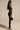 A woman with long hair stands in profile against a neutral backdrop, exuding vintage romance. She wears the WISH YOU WERE ME LACE JUMPSUIT with a plunging neckline, paired with a small black handbag and black high-heeled sandals to complete her elegant look.