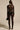 The rear view shows a person in the WISH YOU WERE ME LACE JUMPSUIT, featuring sheer black lace with long sleeves and pants, radiating vintage romance. They accessorize with high heels and a black handbag, standout against the plain light-colored studio backdrop.
