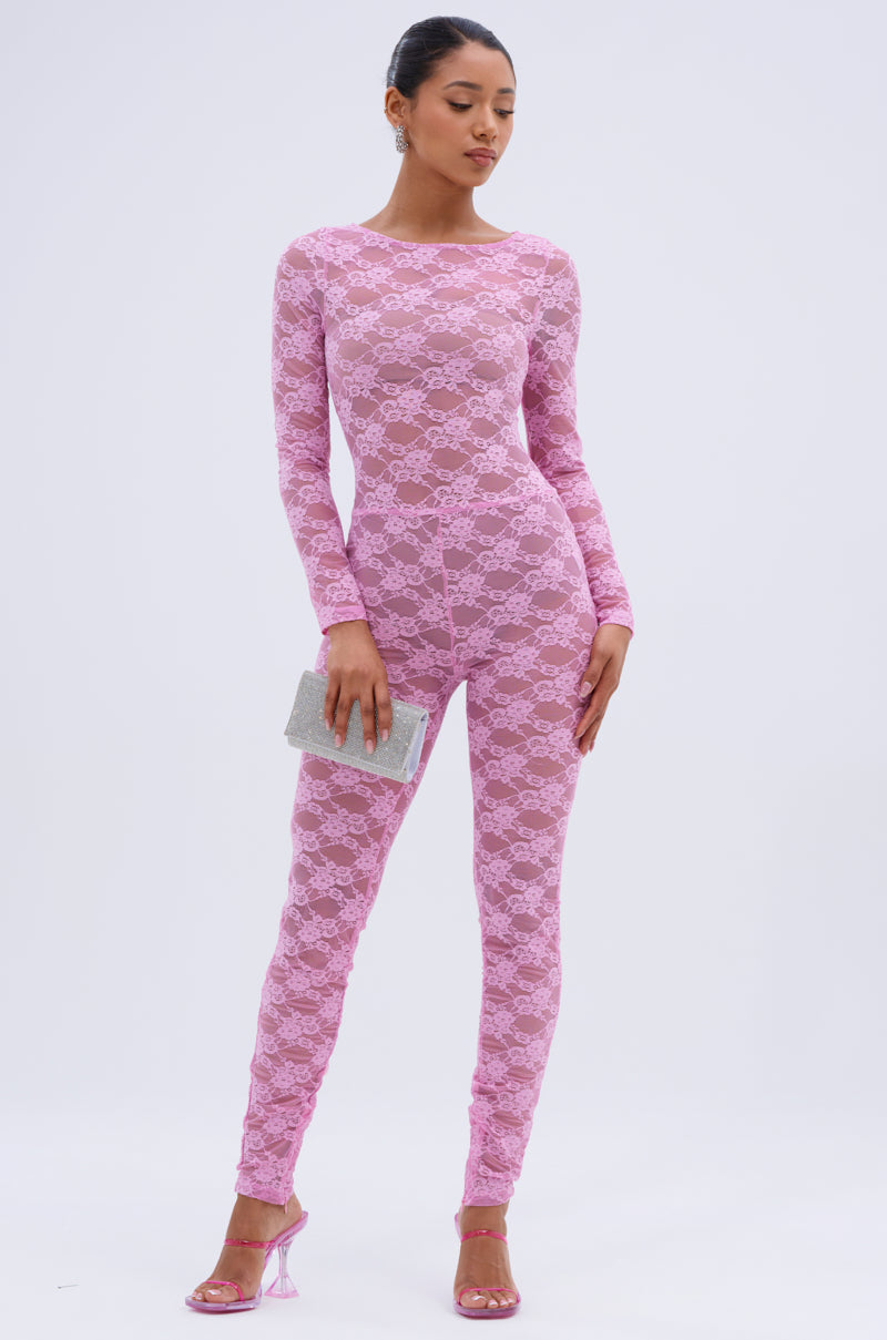 A woman stands confidently against a white background in the FROM DAY TO NIGHT LACE JUMPSUIT IN LIGHT PINK, featuring fitted long sleeves and floral lace, paired with pink heels and a silver clutch. Her dark hair is styled in a neat bun.