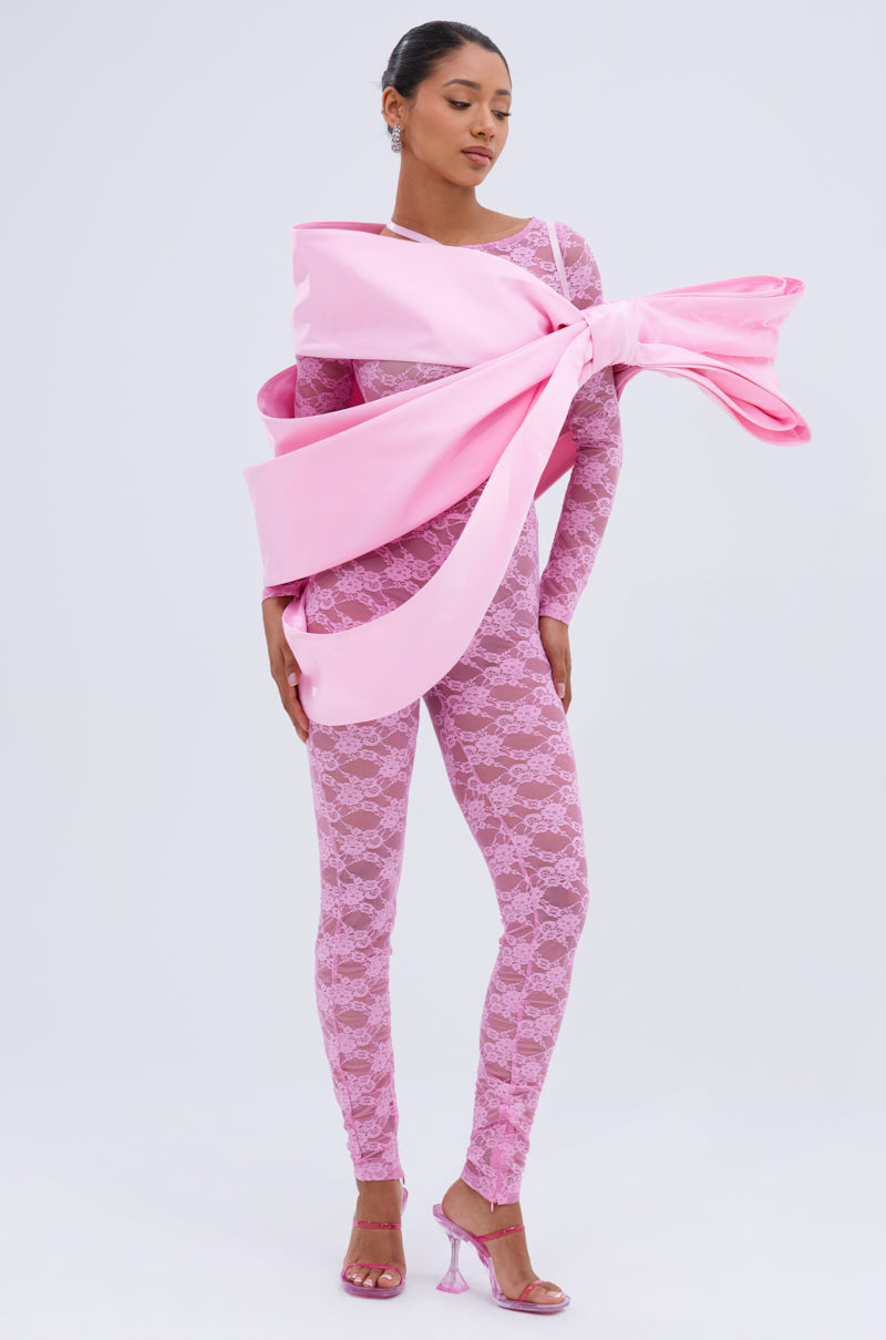 A woman wears the FROM DAY TO NIGHT LACE JUMPSUIT IN LIGHT PINK with pink heels and an oversized dramatic bow wrapped around her torso, standing against a plain white background.