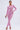A woman poses confidently in the FROM DAY TO NIGHT LACE JUMPSUIT IN LIGHT PINK, featuring fitted long sleeves and a pink floral lace pattern. She accessorizes with a silver clutch and pink heels—ideal for day-to-night style.