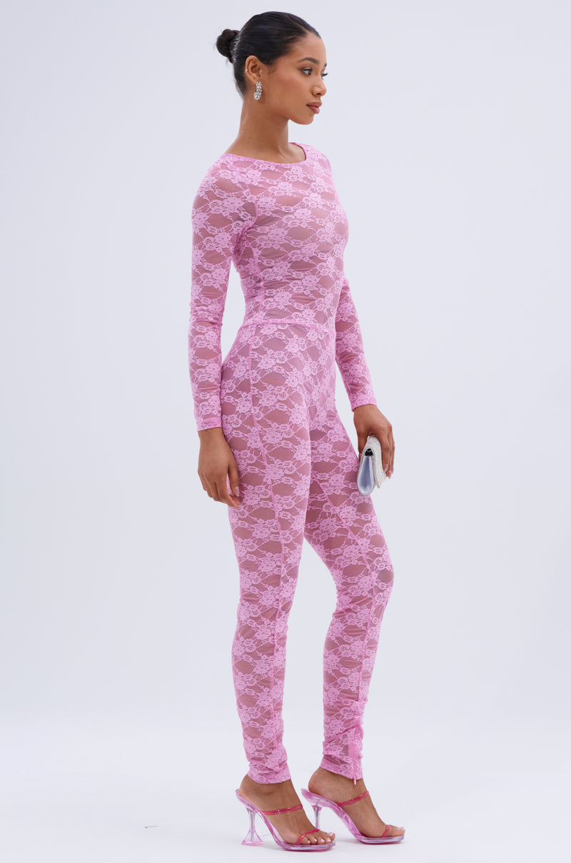 A woman stands sideways in the FROM DAY TO NIGHT LACE JUMPSUIT IN LIGHT PINK with matching pink heeled sandals, a small silver clutch, and earrings. Her hair is styled in a neat bun.