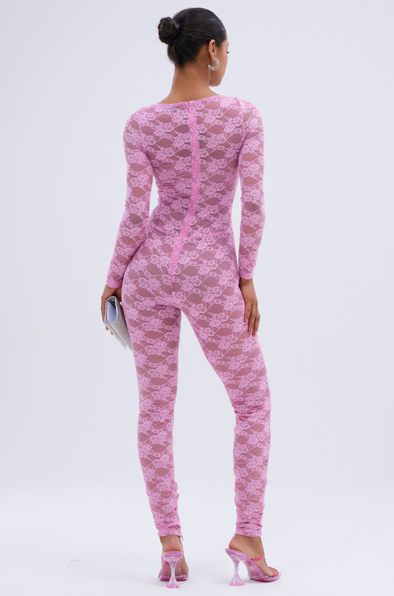 A woman faces away in the FROM DAY TO NIGHT LACE JUMPSUIT IN LIGHT PINK, a fitted, long-sleeve floral lace style. She pairs it with pink heeled sandals, a small clutch, and a neat bun for a chic day-to-night look.