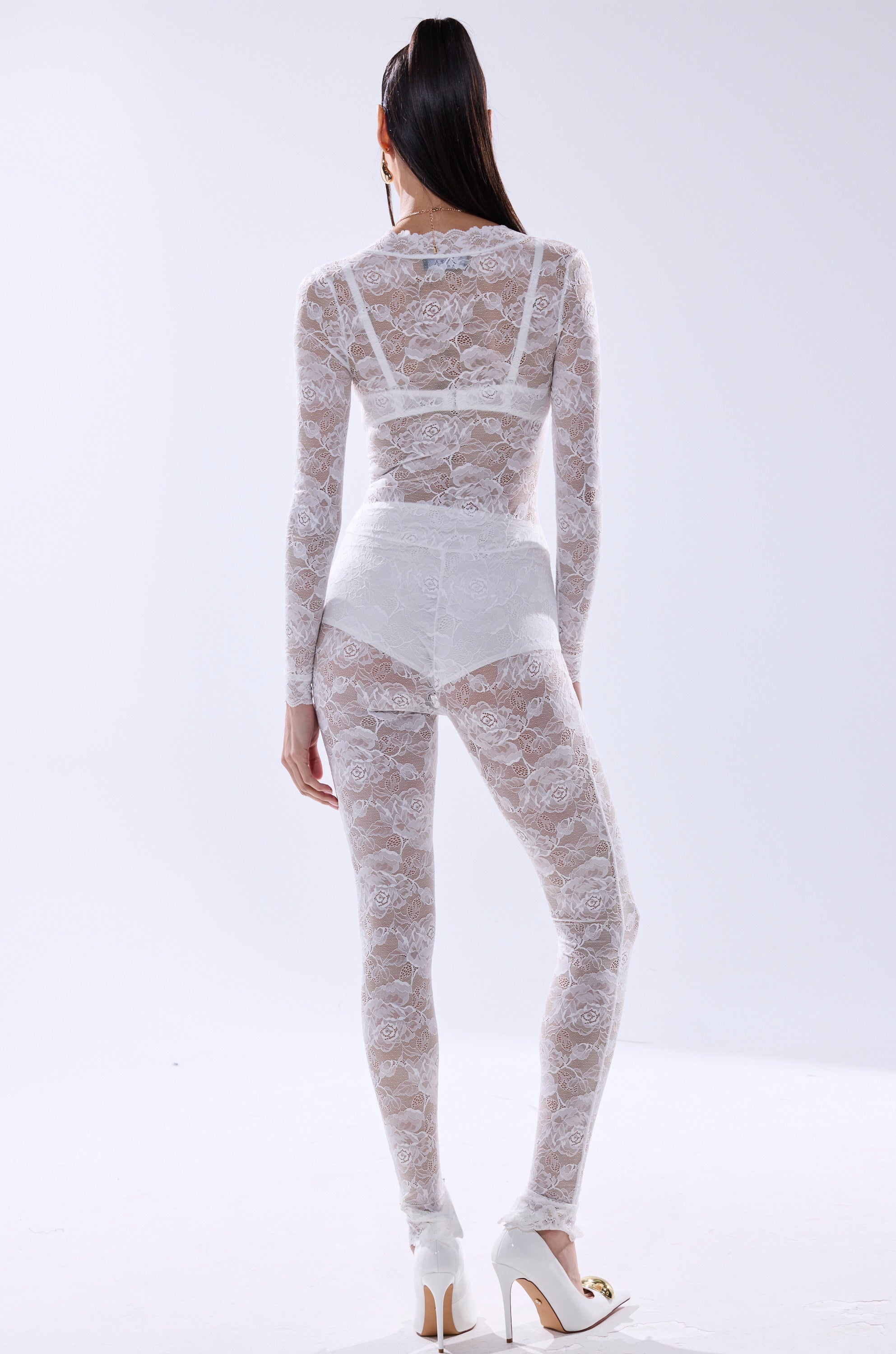 A person with long dark hair in a ponytail wears the WISH YOU WERE ME LACE JUMPSUIT IN WHITE—sheer, long-sleeved, and paired with matching high-waisted bottoms and white heels—facing away against a plain, light background.