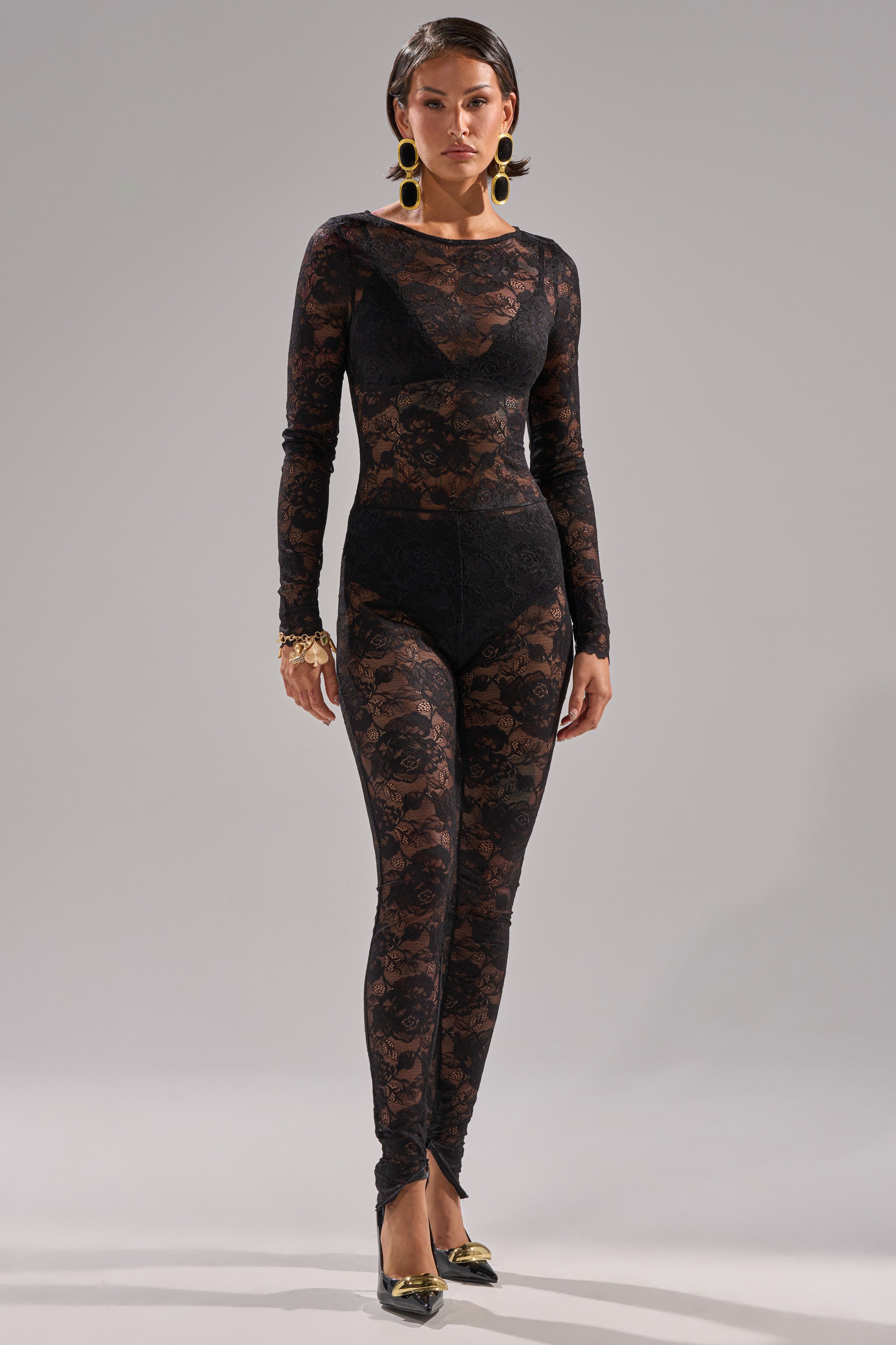 A woman poses against a plain background in the FROM DAY TO NIGHT LACE JUMPSUIT IN BLACK, styled with high-waisted underwear, large gold earrings, a bracelet, and black pointed heels featuring gold embellishments.