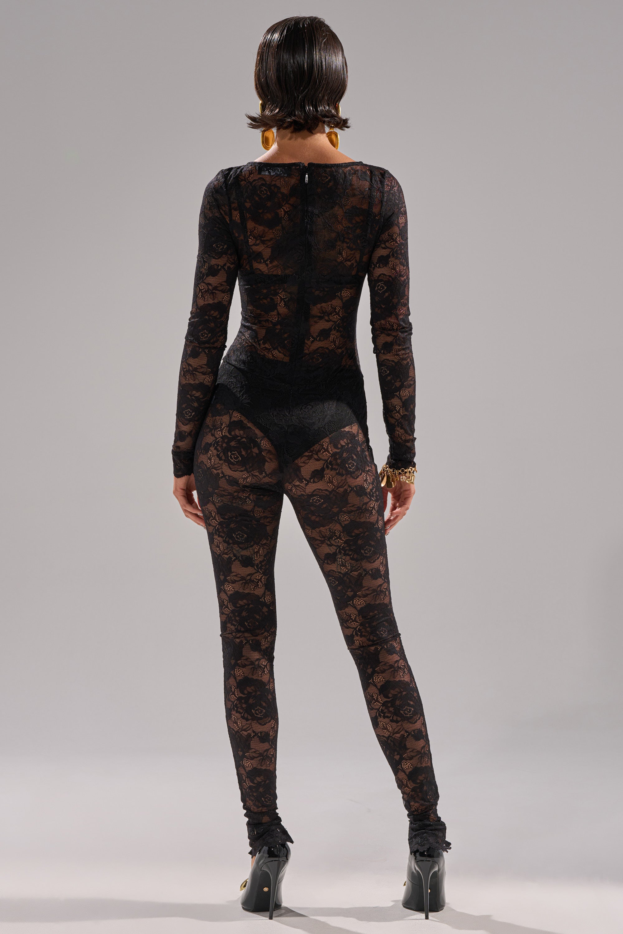 A person faces away, wearing the FROM DAY TO NIGHT LACE JUMPSUIT IN BLACK—a sheer, long-sleeve lace one-piece with leggings—paired with black heels, gold accessories, and slicked-back hair. The background is plain light gray.