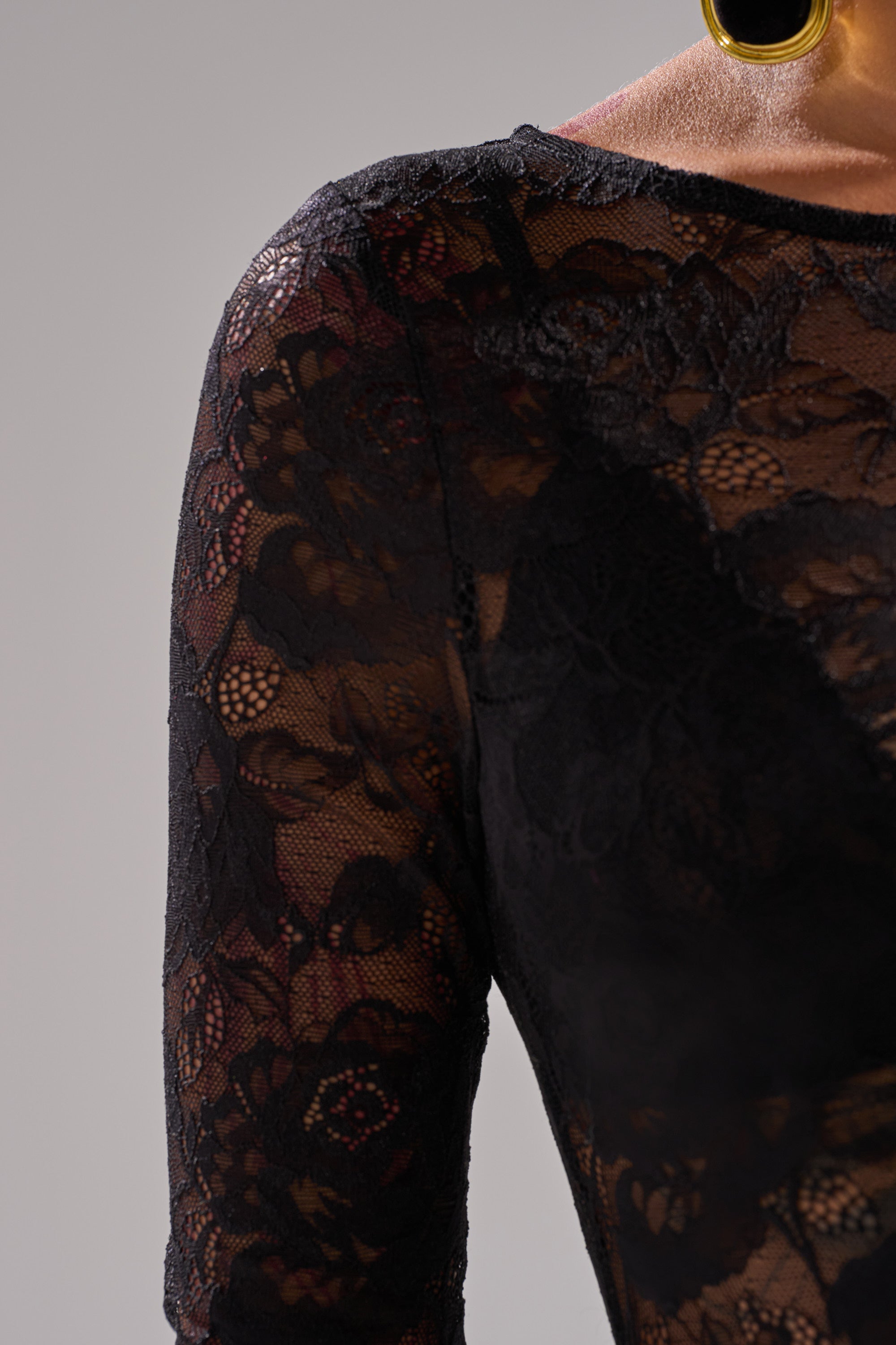 Close-up of a person with dark skin wearing the FROM DAY TO NIGHT LACE JUMPSUIT IN BLACK, featuring sheer black lace with floral patterns and large round gold earrings, set against a plain light gray background.