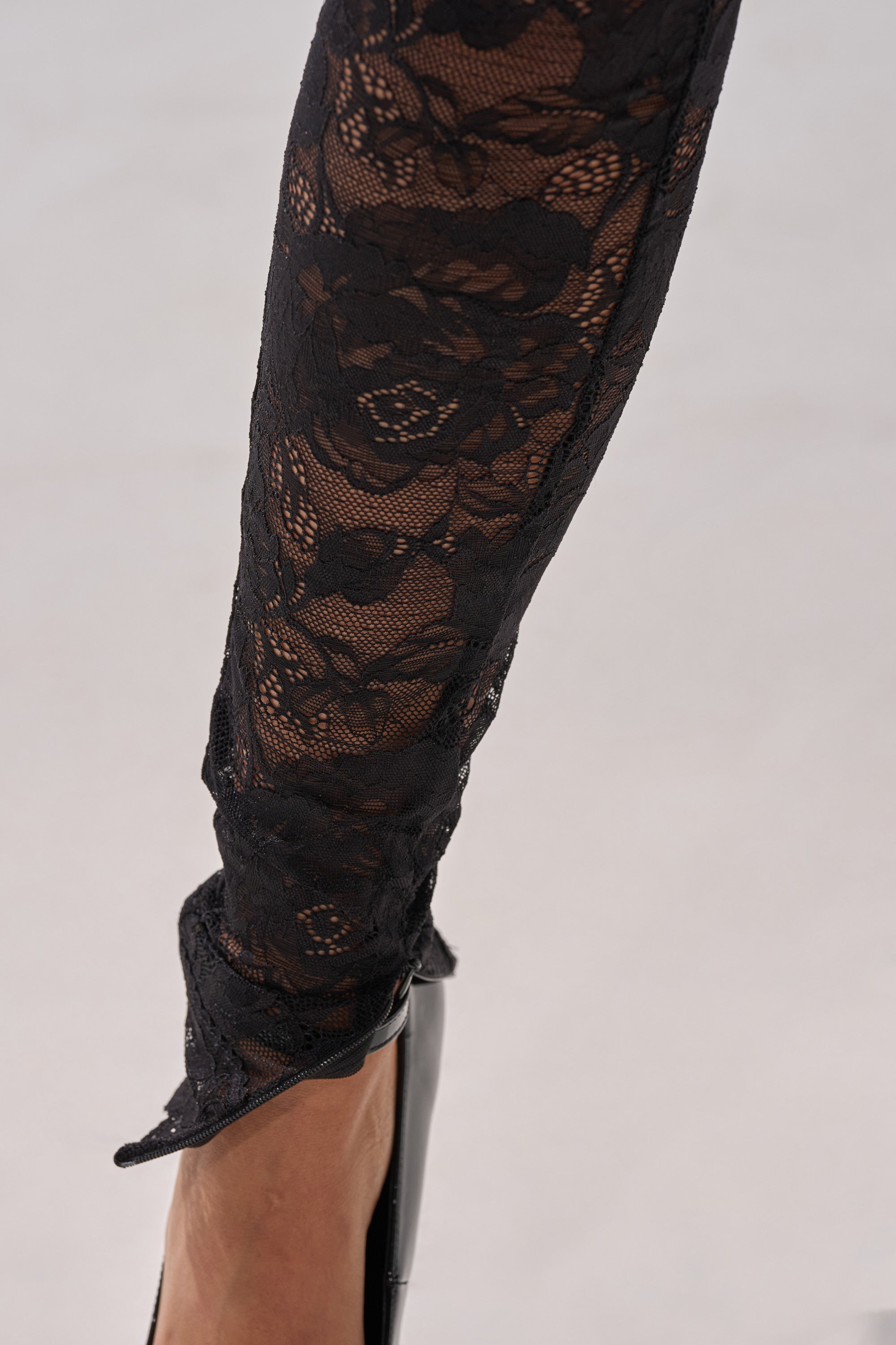 A close-up of a lower leg in black lace tights and a pointed-toe shoe, paired with the FROM DAY TO NIGHT LACE JUMPSUIT IN BLACK, set against a neutral background.
