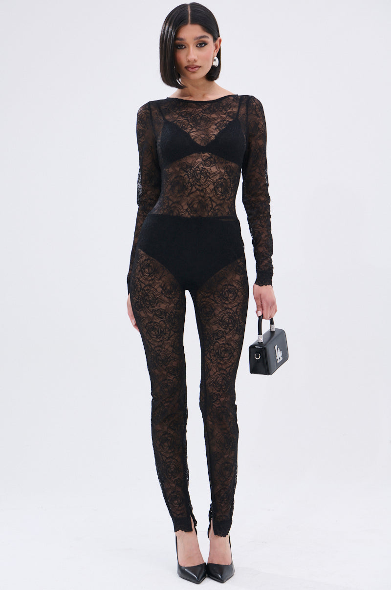 A woman with straight dark hair and a neutral expression stands against a plain background, wearing the ONLY IF THERE'S BOTTLE SERVICE LACE JUMPSUIT over black undergarments, paired with black heels and a small black purse.