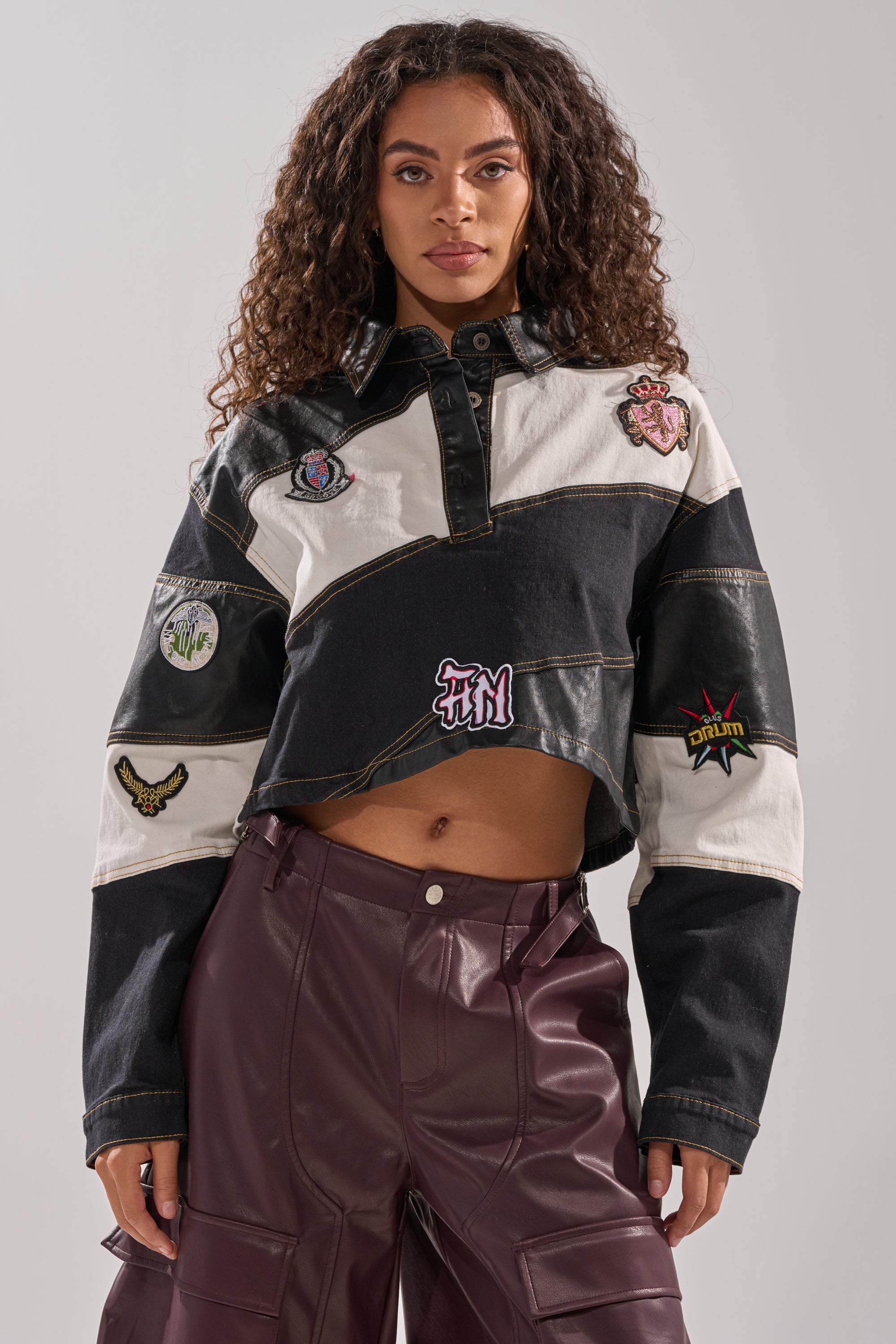 A woman with curly hair wears the GUESS WHAT STRIPE COLLARED BLOUSE, styled with high-waisted, burgundy leather pants, and stands against a plain background.