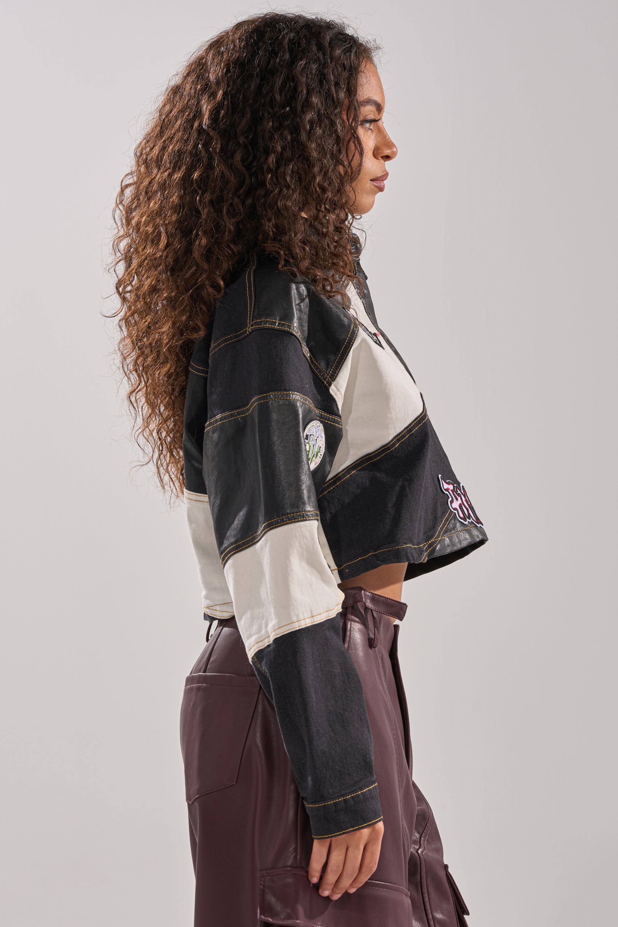 A woman with long curly hair stands in profile, wearing the GUESS WHAT STRIPE COLLARED BLOUSE and high-waisted burgundy pants against a plain light background.