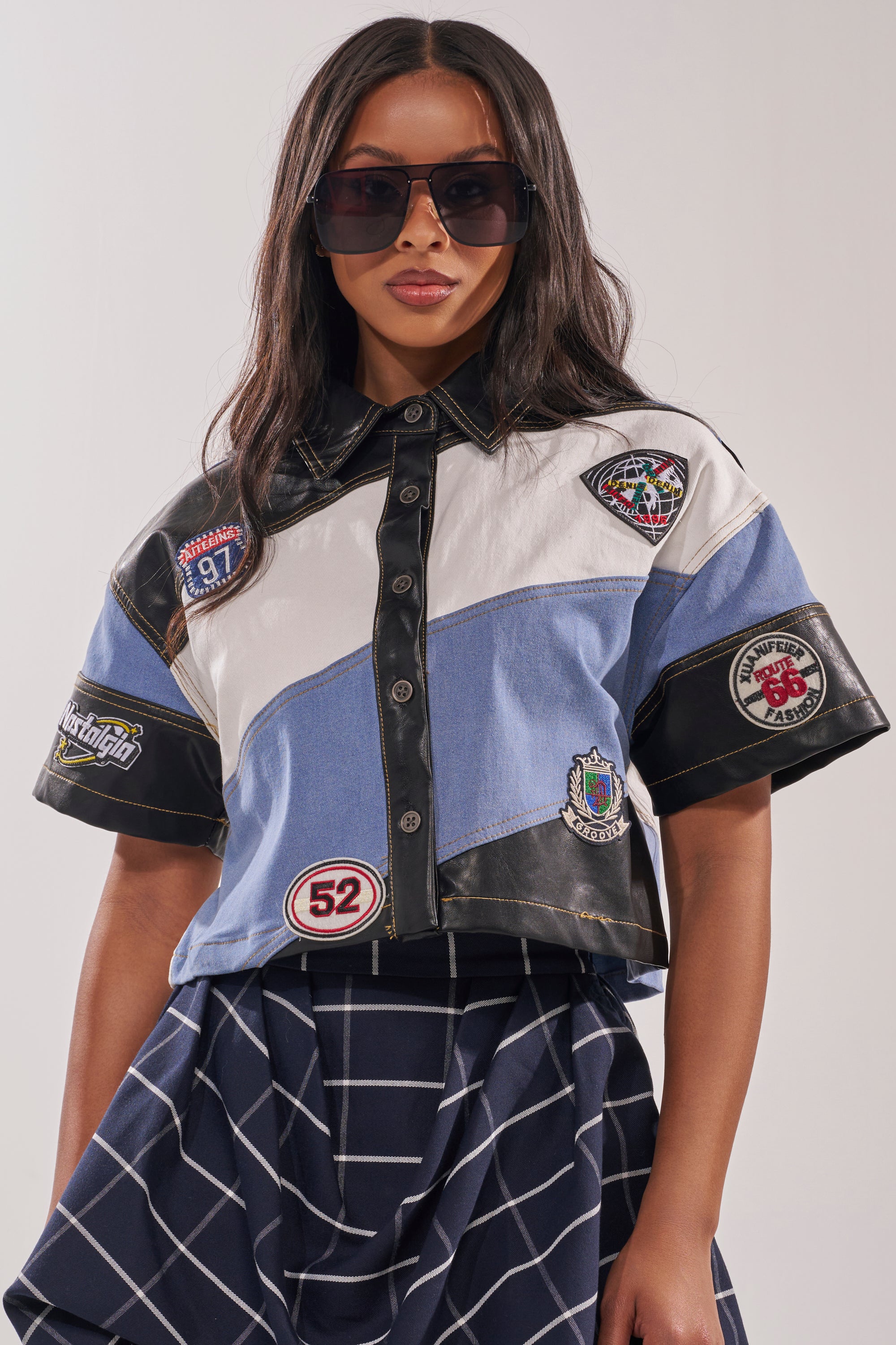 A woman wearing oversized sunglasses and the GUESS WHAT STRIPE SHORT SLEEVE BUTTON DOWN stands against a plain background, styled with a navy plaid skirt and motorsport-inspired patches.
