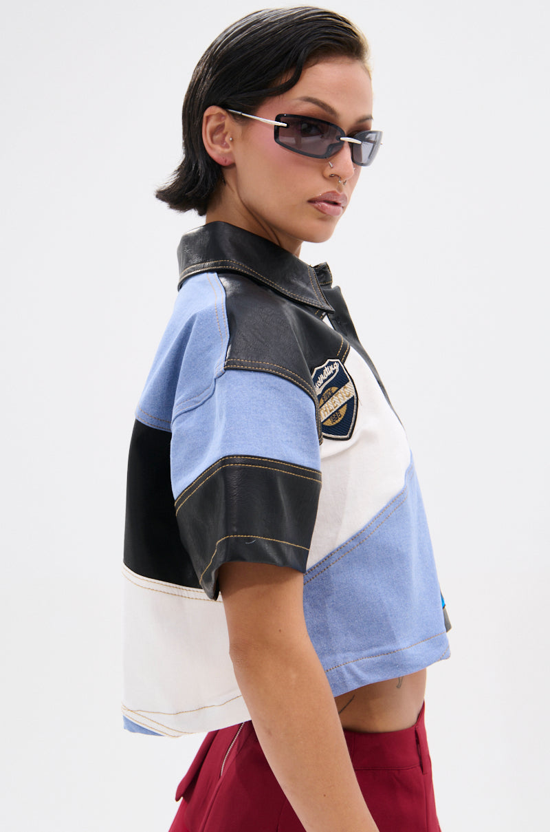 A person with short dark hair and sunglasses poses against a plain light background, wearing red pants and the GUESS WHAT STRIPE COLLARED BLOUSE—a color-blocked, blue, black, and white cropped shirt with a chest patch for streetwear flair.