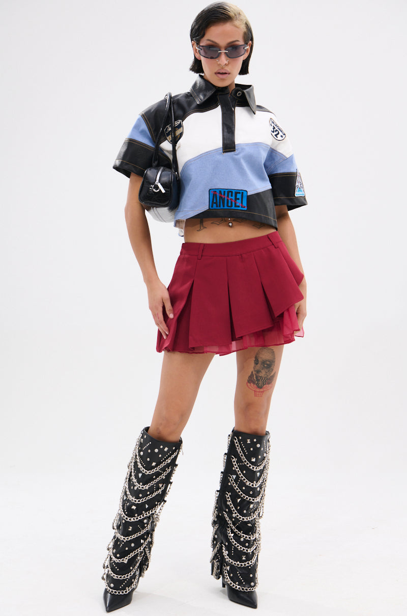 A person wearing sunglasses, the GUESS WHAT STRIPE COLLARED BLOUSE, a red pleated mini skirt, and tall black chain-embellished boots stands confidently against a plain white background.