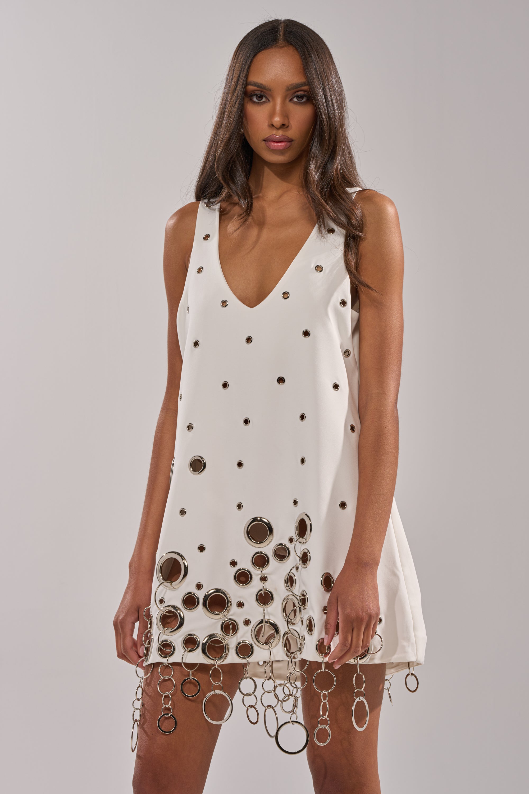 A woman stands against a neutral background wearing the ONE OF A KIND MINI DRESS WITH METAL HARDWARE IN WHITE, featuring a deep V-neckline and metallic grommet and ring accents near the hem.